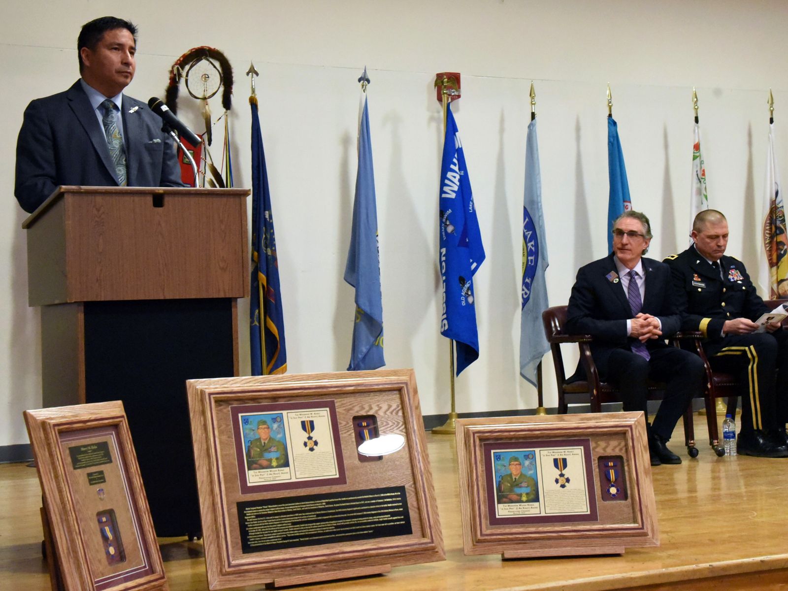 Scott Davis, in his capacity as the executive director of the North Dakota Indian Affairs Commission, speaks at the Circle of Nations/Wahpeton Indian School in Wahpeton, North Dakota, at an awards ceremony honoring North Dakota Army National Guard members who performed acts of courage and selflessness. Then-governor Doug Burgum sits nearby.
