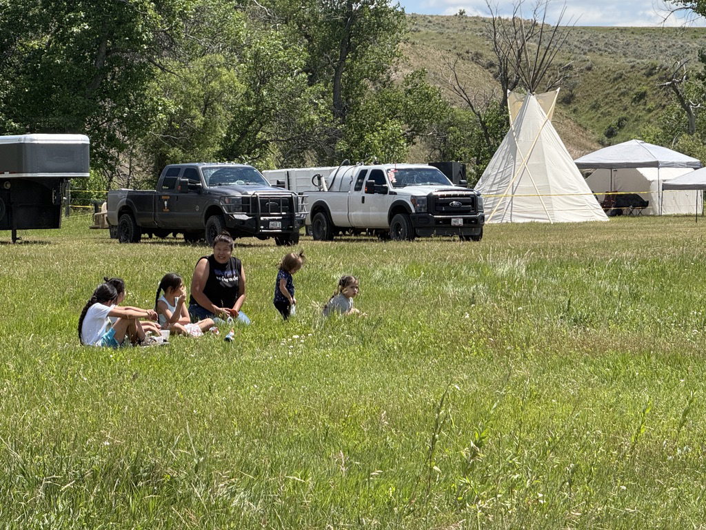 Attendees, including families and young children, are taking time to rest in the grass near the encampment site.