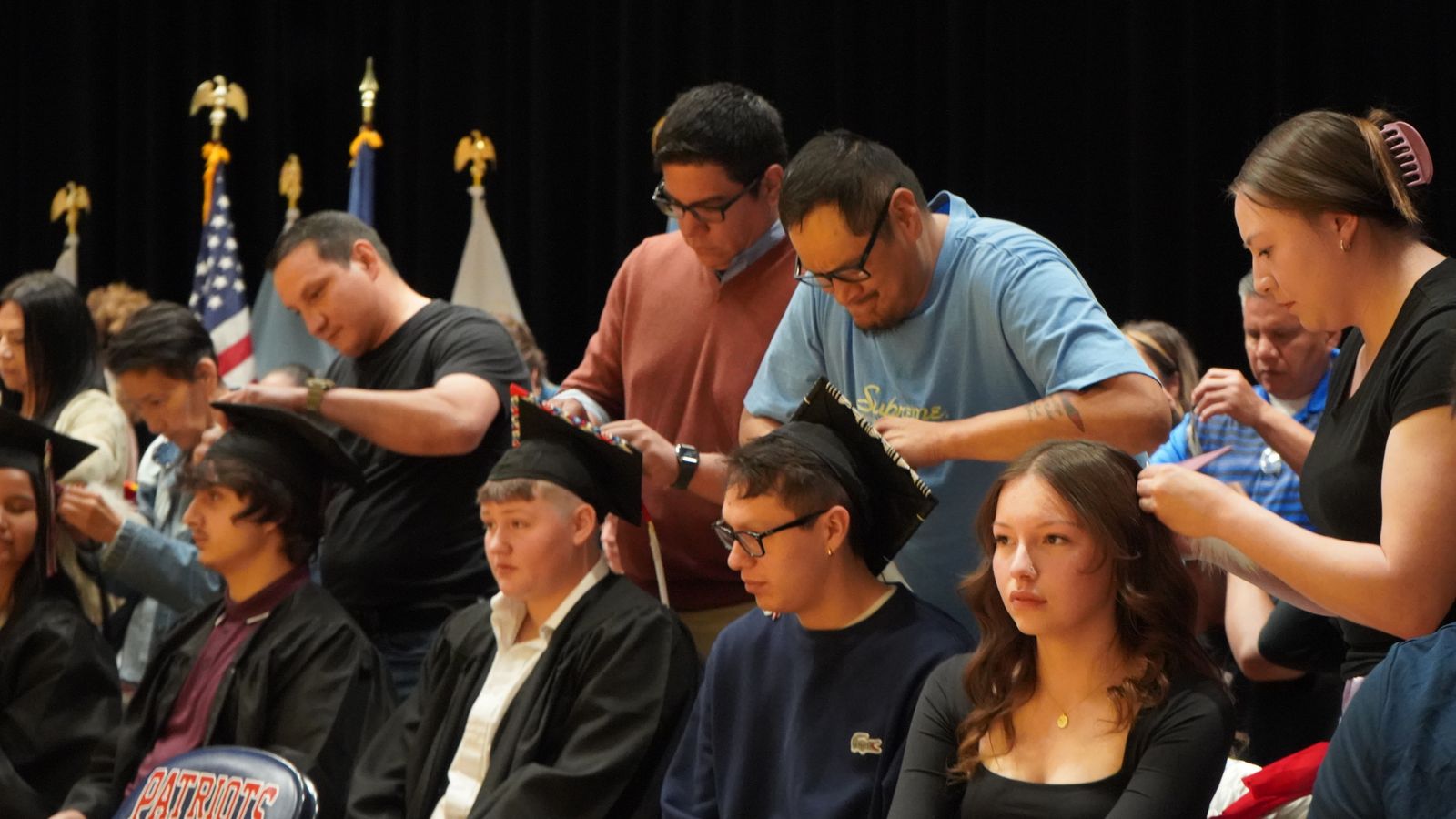 Mandan High School seniors Katawna Bailey, Marle Baker, Jerzi Brugh (right to left) sit as family members tie feathers into hair and onto graduation caps on May 15, 2024. . Blair Baker ties a good knot on his son Marle’s mortarboard.(Photo Credit, Jodi Rave Spotted Bear).
